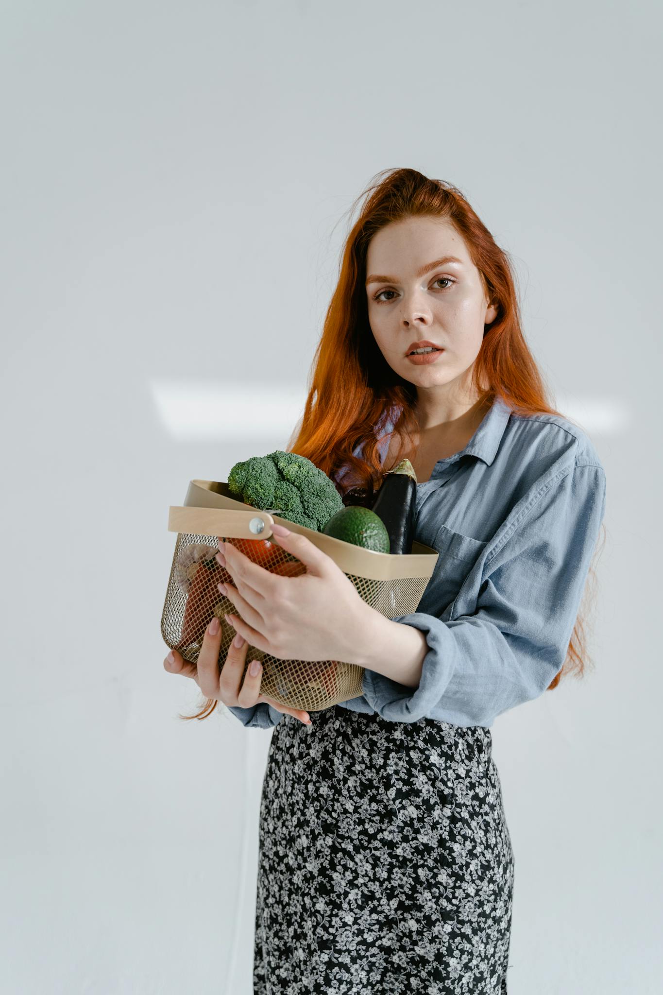 A young woman with red hair holds a basket of fresh vegetables, including broccoli, indoors.